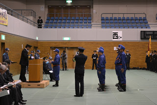 演題に立つ制服を着た関係者の方と消防団の制服を着た隊員の代表者が向かい合って立っており、その後方には消防団の制服を着た隊員が二列に並んでいる出初式の様子の写真