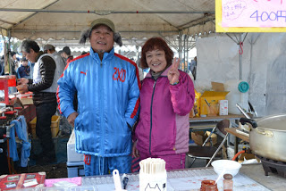 イベント会場の「400円」と書かれたポップが書かれた販売ブースで、青とピンクのジャケットを着た2人がテントの下に立ちピースサインをしている写真