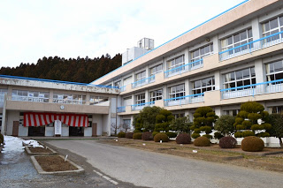 ベージュと白を基調に水色の手すりがある校舎の前に、赤白の幕で飾られた入口と雪が残る整備された庭が写された学校の外観の写真