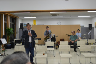 演奏前にスピーチをしている男性が前に立ち、後方にはサックスやドラムを持った演奏者たちが椅子に座って待機している開演前の音楽会の写真