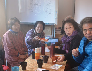 背景には絵や文字が描かれたホワイトボードがある室内で、カップや水筒が置かれたテーブルを囲んで座る4人の人たちがピザを試食し、奥に座っている女性が焼きあがったピザを持っている団らんの様子を写した写真