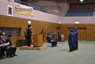 演台に黒い制服を着た関係者が立ち、演題の前に青色の制服姿の4人の団員が一列に整列し出発式の式典が厳かに進行している様子の消防団・交通指導隊出初式の写真