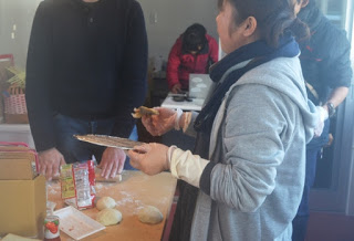 テーブル上には箱や缶、料理や製菓の材料が置かれ、手前に立つ女性が焼いたばかりのピザを試食している様子の写真