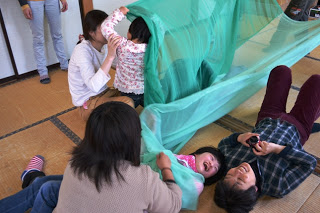 緑色の薄い布をくぐる遊びをしている子どもたちと大人が、一緒に笑い合いながら楽しんでいる様子を写した写真