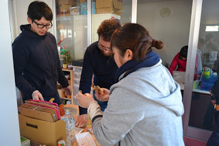 ペットボトルなど様々な物が置かれたテーブルの周りに三人の人物が立ち、出来たばかりのピザを食べている様子の写真