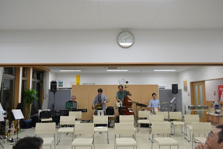 ステージ前に白い椅子が整然と並べられ、その奥で男性4人の演奏者がベースやサックスなどの楽器を構えて立っている演奏準備中の写真