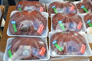 テーブルの上にそれぞれに青・緑・赤の日本語ラベルが貼られている、ビニール包装された複数のタコの足のパックが並べられた商品の写真