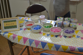 カラフルな模様のテーブルクロスがかけられたテーブルの上に、赤・黄・青・緑・ピンク・紫などの材料が入った白い皿が並べられており、左側には「きゃんどる」と書かれた額入りの看板が置かれたキャンドルづくりコーナーの写真