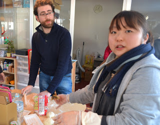 テーブル上にはペットボトル、ラップの箱、食品などが並び、テーブルの前に立つ二人の人物が食品の準備作業をしている様子の写真