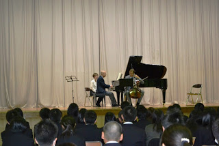 手前で生徒たちが座って演奏を見ているなか、背景には大きなカーテンが広がり、ステージ上でピアノ奏者がグランドピアノを演奏し、もう1人の演奏者がチェロを弾いている写真