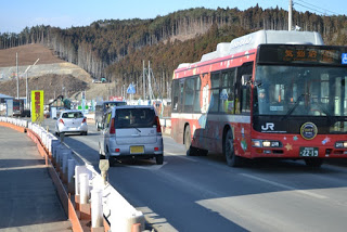 周囲は丘や木々が広がり、遠くには工事用の機材も見え、道路の脇にはバリケードが設置され、目的地が示された赤いJRバスや複数の車両が走る道路の様子の写真