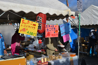 屋外の屋台で、「ちいたこ チーズ入りたこやき 400円」などが書かれた、赤・黄色・青のカラフルなポップが3枚掲げられており、カウンターの後ろではスタッフが準備している、たこ焼き屋台の写真