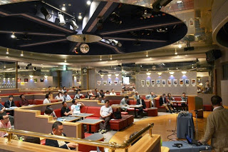 天井に大きな装飾のあるホールで、参加者たちが赤いソファに座って講演を静かに聞いている写真