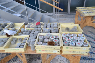 階段の手前に木製のテーブルの上に複数のトレイが並べられ、それぞれ一部には種類や試験内容を示すような色付きの印がある小さな円柱状のコンクリート試料が収められた屋内の写真