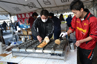 屋外イベントのテントの下で、赤いジャケットを着た男子生徒と黒い服の男性教師が大きな鉄板でおやきを作っている屋台の写真