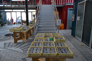 モザイク模様の床が配置された中央に階段があり、その前に円柱状の物体が並べられたトレイが複数置かれたテーブルが並び、左側のガラス扉越しには車両が見える屋内空間の写真