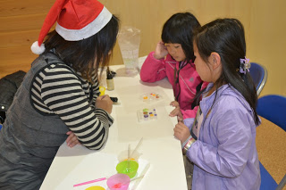 室内でサンタ帽をかぶった女性と、2人の女の子がテーブルに向かい合って座っており、テーブルの上にはカラフルなボタンなどクラフトの材料が置かれているワークショップの様子の写真