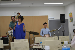 青い服を着た女性がマイクを持って歌っており、その後ろでコントラバスとドラムの演奏者が伴奏している歌唱中の写真