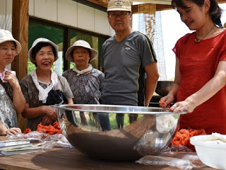 大きなステンレスのボウルが置かれた作業債の周りに女性3人と男性1人が集まり、赤いシャツを着た講師の女性が草木染を実演している様子の写真