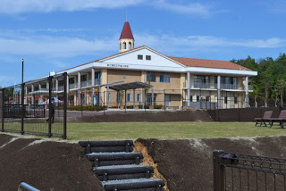 赤い屋根と塔を持つ二階建ての校舎が青空の下に建ち、芝生と階段が手前に広がっている南三陸町立戸倉小学校の外観の写真