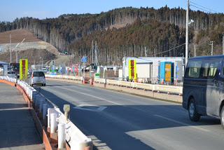 空は晴れていて、背景には木々に覆われた丘と建設現場と思われる建物や構造物が見え、道路沿いには黄色地に赤文字の看板が複数立てられており、白い車や濃い色のバンなどの車両が走る道路の両側に工事用のバリケードが設置された写真