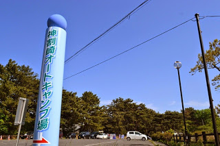 青い空の下、木々に囲まれ複数の車が停まっている駐車場の入り口に、水色で縦長の「神割崎オートキャンプ場」と書かれた看板が立っている写真