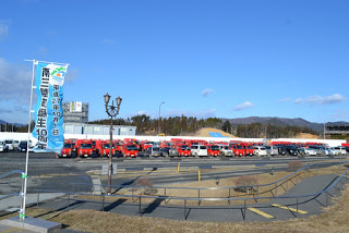 澄んだ青空の下、背景には建物と木々、さらに遠くに山々が広がり、手前には「南三陸町誕生10周年」と書かれた水色の縦長ののぼり旗が立っている敷地内の広い駐車場に多数の赤い消防ポンプ車が整然と並んでいる写真