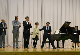 グランドピアノがあるステージ上に、6人の人々がフォーマルな服装で立っていて、何人かは右側から歩いて来る女子生徒を見ながら拍手をしている様子の写真
