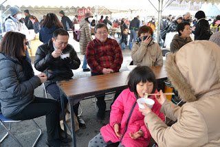 屋外イベントのテントの下で、ピンクのジャケットを着た子どもやベージュのコートを着た大人などがテーブルを囲んで座っており、それぞれ購入した食べ物を食している写真