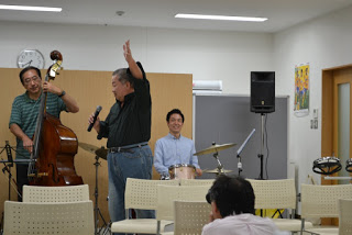 黒いシャツの男性が片手を上げて歌っており、後ろにはベース演奏者と笑顔のドラマーが演奏している音楽会の一幕の写真