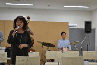 黒い服の女性がマイクを持ってステージの前に立ち、ドラム演奏者とコントラバス奏者が後方で伴奏している歌唱中の写真