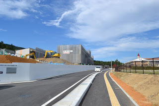 右側に戸倉小学校の校舎、左側に工事中の建物とショベルカーが並び、中央の道路には白い車が走っている風景の写真