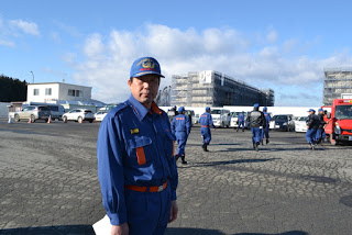 背景には赤い消防車両や建物が見える駐車場で、青い制服を着た元木分団長が立っている様子をアップで撮影した写真