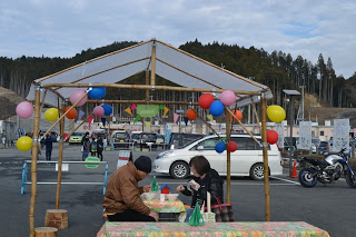 カラフルな風船が飾られた竹の柱と半透明の屋根で作られたテント状の屋外構造物の下に置かれたテーブル席に座る男女が食事をしている様子の写真