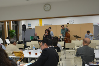 数名の聴衆が座っている中、ステージ上ではキーボード、コントラバス、ドラムの演奏者に加え、中央に立つ女性ボーカルが歌っている演奏中の写真