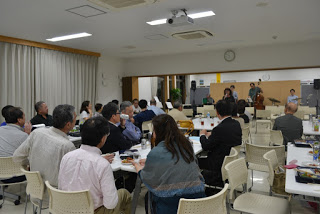 テーブルを囲む多くの観客が飲み物を手にしながら演奏を見つめており、ステージではバンドとボーカルが音楽を披露している音楽イベント会場の写真