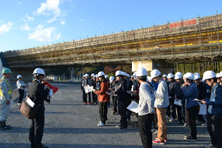 建設中の高架橋を背に、ヘルメットをかぶった見学者たちが整列して説明を受けている写真