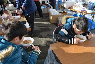 屋外に設置された販売ブースのテントの隅で、2人の男の子がテーブルに座って温かい麺類を食している様子の写真