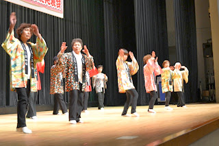 舞台上でカラフルな和柄の法被を着た方々が、両手を上げて踊りを踊っている様子の写真