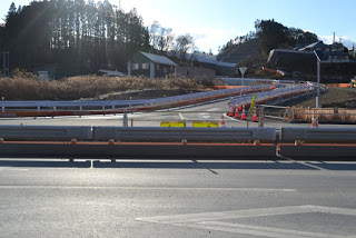 空は晴れて雲が少し浮かんでいる背景には木々や建物、丘が見え、複数車線の道路で、オレンジと白のバリケードや三角コーンによって一部の車線が封鎖されている工事現場の様子が写された写真