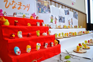 赤い毛氈の上に小さな色とりどりの人形が並べられ、「ひなまつり」と書かれた紙が掲示されている室内展示の写真