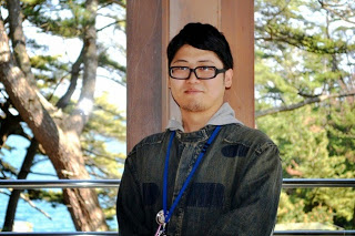 太陽の光が反射した木々を背景に、施設の柱の前に立つメガネをかけた山田虹太郎さんをアップで撮影した写真