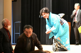 舞台の上で青い着物を着た女性が中腰になり、舞台下の男性達からおひねりをもらい話をしている様子の写真