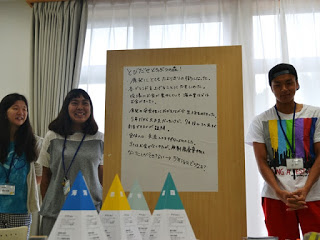 模造紙にまとめた意見を背景に、3名の参加者が前に立って発表を行っており、テーブルの上にはボードゲームのコマが並んでいる発表の写真