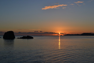 地平線に沈みかけた太陽が空と水面をオレンジ色に染め、雲がわずかに浮かぶ中、手前に大きな岩または小さな島が二つ見える穏やかで美しい初日の出の様子の写真