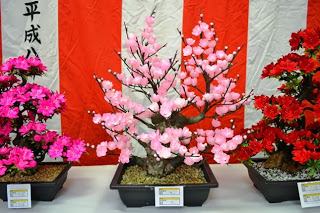 紅白幕の前に、モモやツツジを模したようなピンクや赤のシルクフラワーが展示されている写真