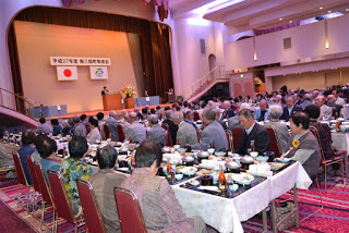 華やかな会場で、多くの高齢者が整然と並べられた食事の席に座り、ステージ上の挨拶を聞いている全体の様子の写真