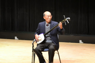 黒いスーツ姿の男性が舞台の中央で椅子に座り、真剣な表情で三味線を演奏している写真