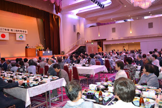 大きな宴会場でテーブルに整然と料理が並び、高齢者たちが着席して式典の話を聞いている様子で、舞台では挨拶が続いている写真