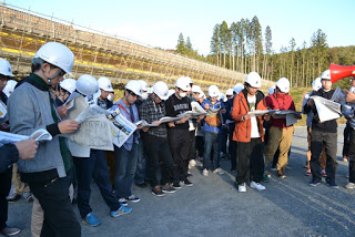 工事現場でヘルメットをかぶった見学者たちが、資料を手に説明を真剣に読んでいる写真